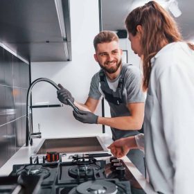 young-professional-plumber-in-grey-uniform-shows-results-of-work-for-housewife-on-the-kitchen-q25w1nopk1bplfeakirhyps008xh221aihxs0o6700